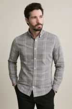 Man with a beard wearing a grey full-sleeve shirt with a white windowpane check pattern, standing with hands in the pockets of his black trousers.