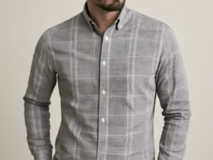 Man with a beard wearing a grey full-sleeve shirt with a white windowpane check pattern, standing with hands in the pockets of his black trousers.