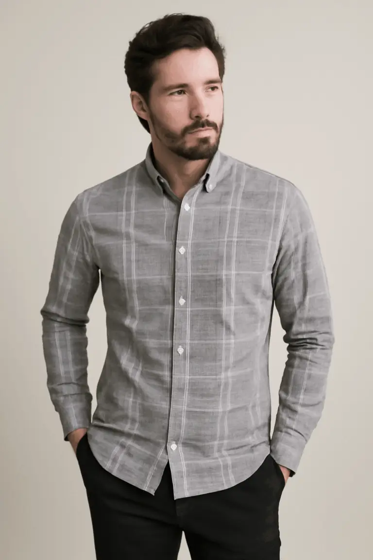 Man with a beard wearing a grey full-sleeve shirt with a white windowpane check pattern, standing with hands in the pockets of his black trousers.