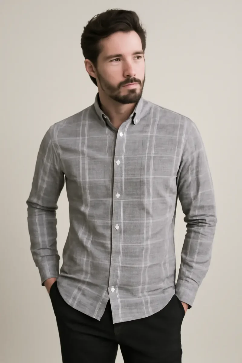 Man with a beard wearing a grey full-sleeve shirt with a white windowpane check pattern, standing with hands in the pockets of his black trousers.